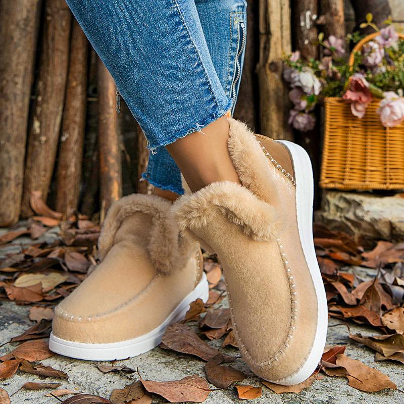 Beige slippers with fur trim worn by a person in blue jeans, standing on a ground with leaves and flowers.