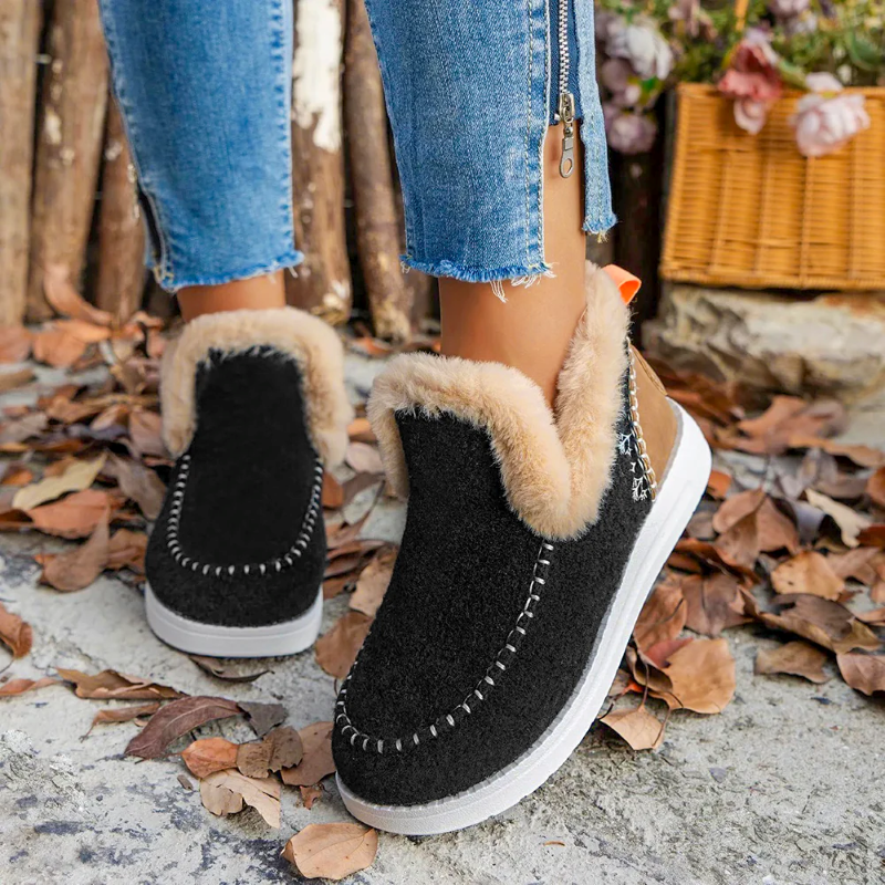 Black slip-on shoes with beige fur lining worn by a person on a leaf-covered ground.
