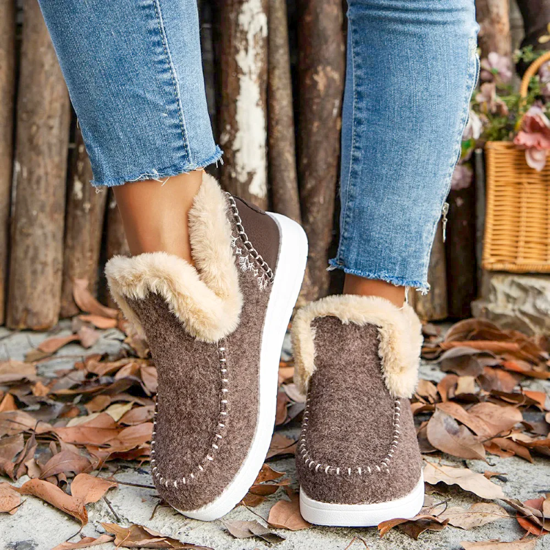 Brown slippers with fur lining worn by a person standing on a leaf-covered ground.
