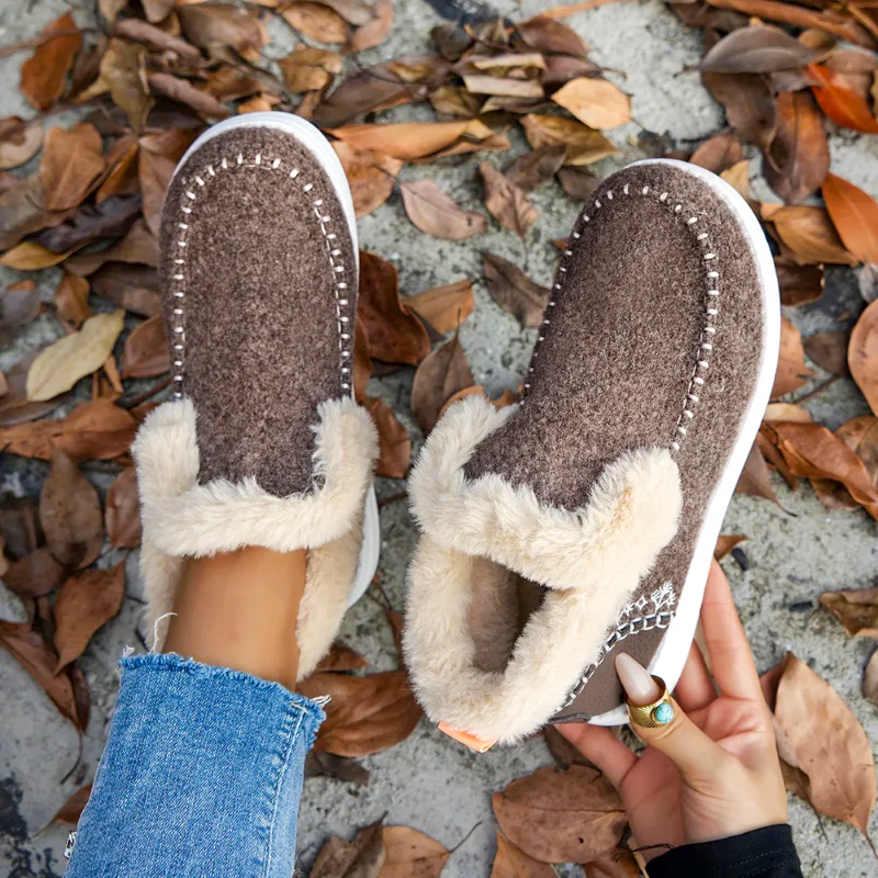 Brown and beige slippers with fur lining on a ground covered with fallen leaves.